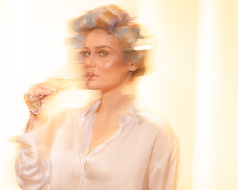Estetska, blago zamućena fotografija plave žene sa viklerima koja pije šampanjac u svilenoj košulji.